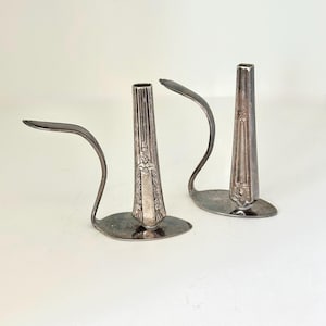 May include: Two silver candle holders. Each holder is made from a repurposed fork, with the tines forming the candle cup and the handle bent into a curved arm. The bases are round and the candle cups are decorated with floral patterns.