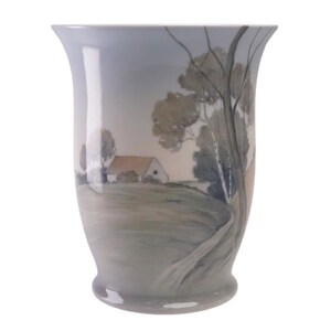 May include: A light blue vase with a landscape painting. The artwork depicts a house, trees, and a field in muted tones of gray, green, and brown. The vase has a flared top and a rounded base.