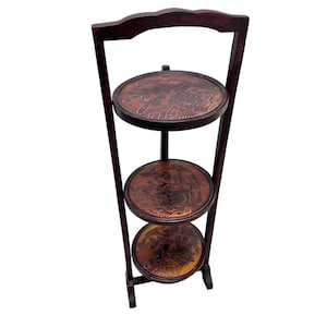 May include: A dark brown, three-tiered serving stand with carved circular shelves. The stand has a handle at the top and a sturdy base. Each shelf features intricate carvings, adding a decorative element to the functional design.
