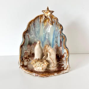 May include: A ceramic nativity scene sculpture featuring a glossy, arched backdrop with blue, brown, and white marbled glaze. A gold-toned star sits at the top. Three abstract figures in cream-colored ceramic are positioned in the foreground.