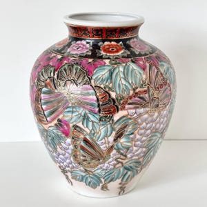 May include: A ceramic vase featuring a detailed pattern of colorful butterflies, green leaves, and clusters of grapes. The neck is decorated with a black band containing floral motifs and a red border with gold geometric accents on a white base.
