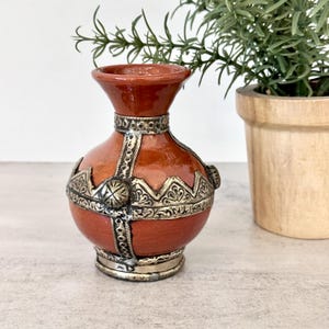 May include: A small, round, terracotta-colored ceramic vase featuring a flared rim and a narrow neck. The vase is decorated with intricate, embossed silver-toned metal overlays that form a cross pattern and a jagged band around the center.