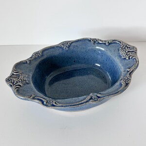 May include: A blue ceramic serving bowl with a speckled finish and ornate, raised floral detailing around the rim. The bowl has an oval shape and a slightly textured surface, suitable for serving food or as a decorative piece.