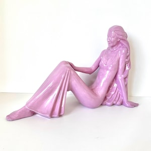 May include: A ceramic sculpture of a seated figure in a flowing gown. The figure is a soft pink color and is posed with one leg extended and the other bent. The sculpture has long hair and is resting on a white surface.