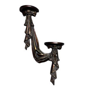 May include: A bronze-colored wall sconce with two circular platforms connected by draped fabric. Each platform is adorned with a bow and tassel details. The sconce has a decorative, ornate design.