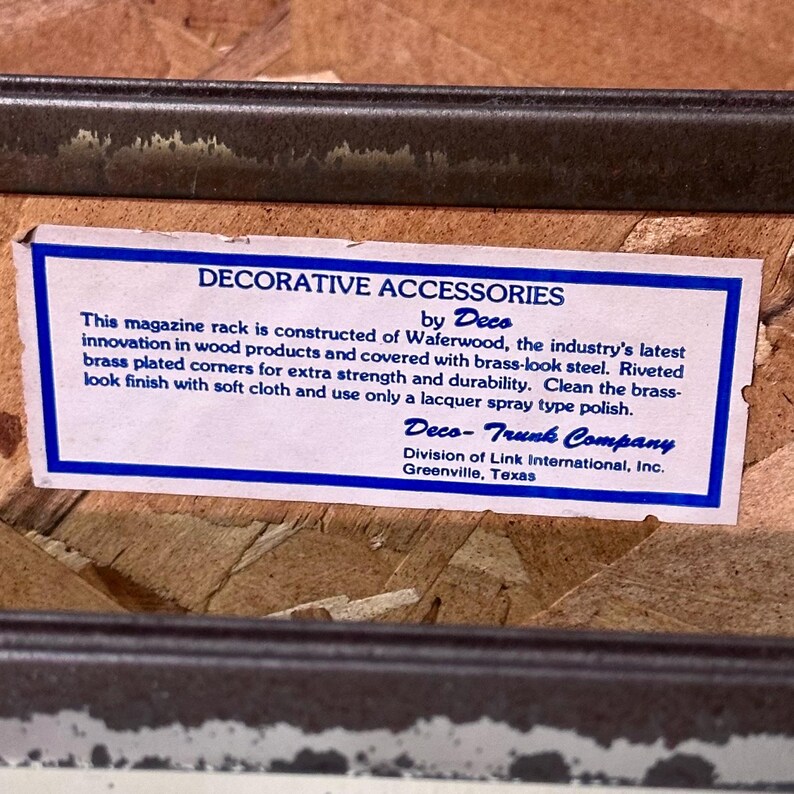 Op de afbeelding: Een tijdschriftenrek met een stalen frame met messinglook en geklonken, messingkleurige hoeken. Het rek is gemaakt van Waferwood en heeft een label met de tekst "DECORATIVE ACCESSORIES by Deco". Het label bevat ook de informatie van Deco-Trunk Company.