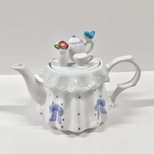May include: White ceramic teapot with a decorative design. The teapot features a white lid with a small teapot, teacup, and a vase with flowers. The body of the teapot is white with blue bows and polka dots. A blue bird is on the handle.