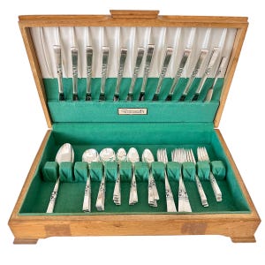 May include: A wooden flatware chest with a hinged lid, lined with white satin and green felt. The interior contains silver-toned utensils, including spoons, forks, and knives, arranged in slots. A small plaque labeled Community is centered inside.