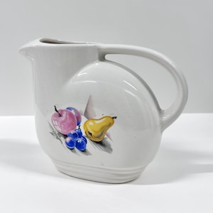 May include: White ceramic pitcher with a curved handle and spout. The pitcher features a painted design of a pink apple, purple plums, yellow pear, and blue grapes. The pitcher is a vintage style.