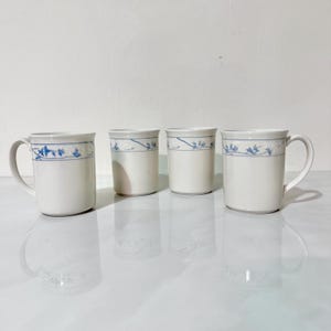May include: Four white ceramic mugs and tumblers with a blue floral design around the rim. The mugs have handles, while the tumblers are handle-less. The set is displayed on a reflective surface.