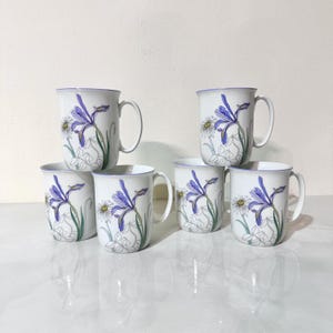 May include: Set of six white ceramic mugs. Each mug features a blue iris and daisy floral design with green stems and leaves. The rims are trimmed with a thin blue line. The mugs have a classic handle design.