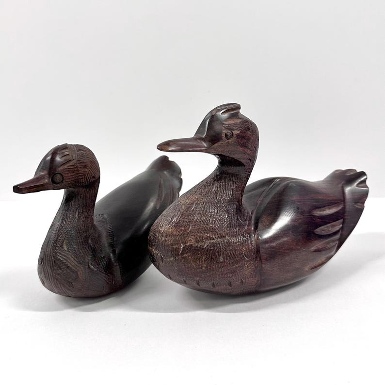 May include: Two hand-carved wooden duck figurines. The ducks are dark brown with detailed feather carvings. One duck is slightly smaller and positioned behind the other. The ducks are displayed on a white surface.