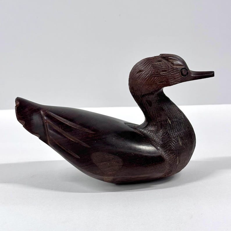 May include: A carved wooden duck figurine in a dark brown hue. The duck is depicted in a resting position, with detailed carving on the head and wings. The beak is long and pointed. The figurine is a decorative item.