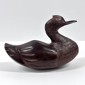 May include: A carved wooden duck figurine with a dark brown finish. The duck has a long neck, a detailed beak, and carved feather details. The figurine is a decorative item, likely for display.