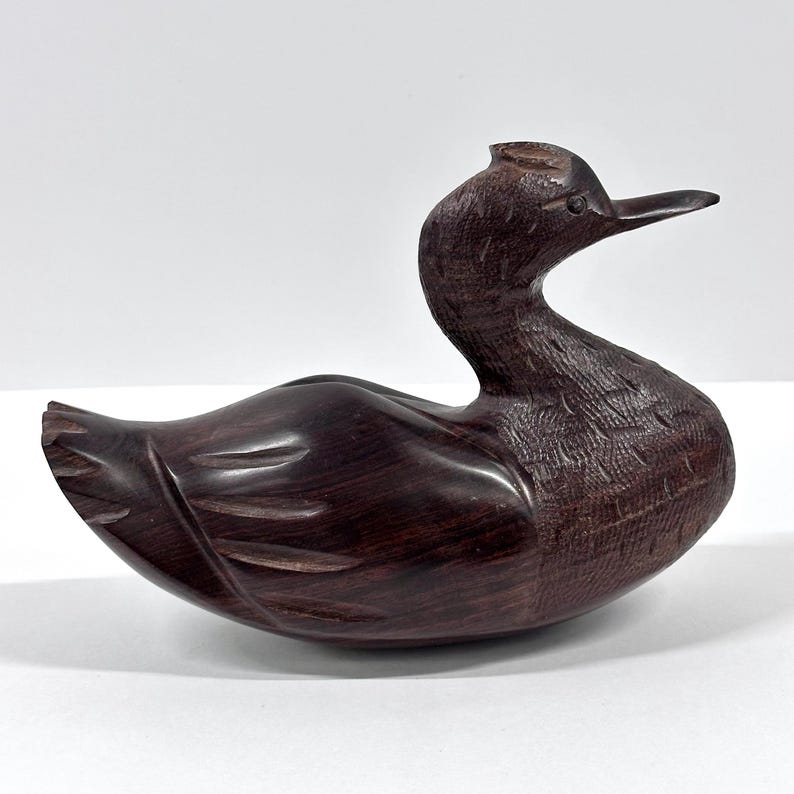 May include: A carved wooden duck figurine with a dark brown finish. The duck has a long neck, a detailed beak, and carved feather details. The figurine is a decorative item, likely for display.