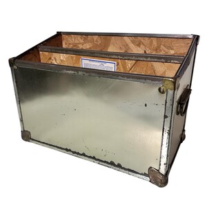 Op de afbeelding: Een rechthoekige metalen opbergbak met een verdeeld interieur. De bak heeft een zilveren buitenkant met messing hoekaccenten en een handvat aan de zijkant. De binnenkant is van hout. Een klein label is binnenin zichtbaar.