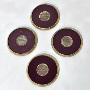 May include: Four round coasters with a burgundy center and a gold-colored rim. Each coaster has a small, gold-colored circle in the center with the text "MICRO 100". The coasters are arranged in a circular pattern.