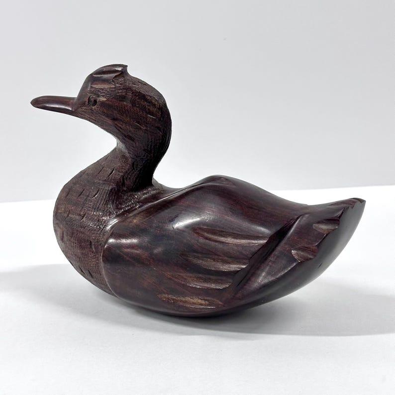 May include: A carved wooden duck figurine in shades of dark brown. The duck has a long neck, a detailed beak, and carved feather details. The figurine is a decorative item, likely for home decor.