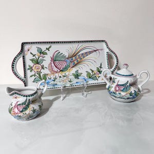May include: A white ceramic serving tray, creamer, and sugar bowl set. The tray has handles and a colorful peacock design. The creamer and sugar bowl also feature the peacock design. The set is decorated with floral and leaf accents.