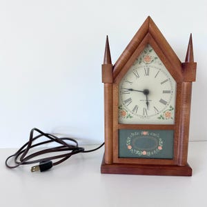 May include: A wooden mantel clock with a pointed gothic arch frame and two decorative spires. The clock face features black Roman numerals, floral accents, and the text Seth Thomas. A dark green panel with a floral border sits below the dial.