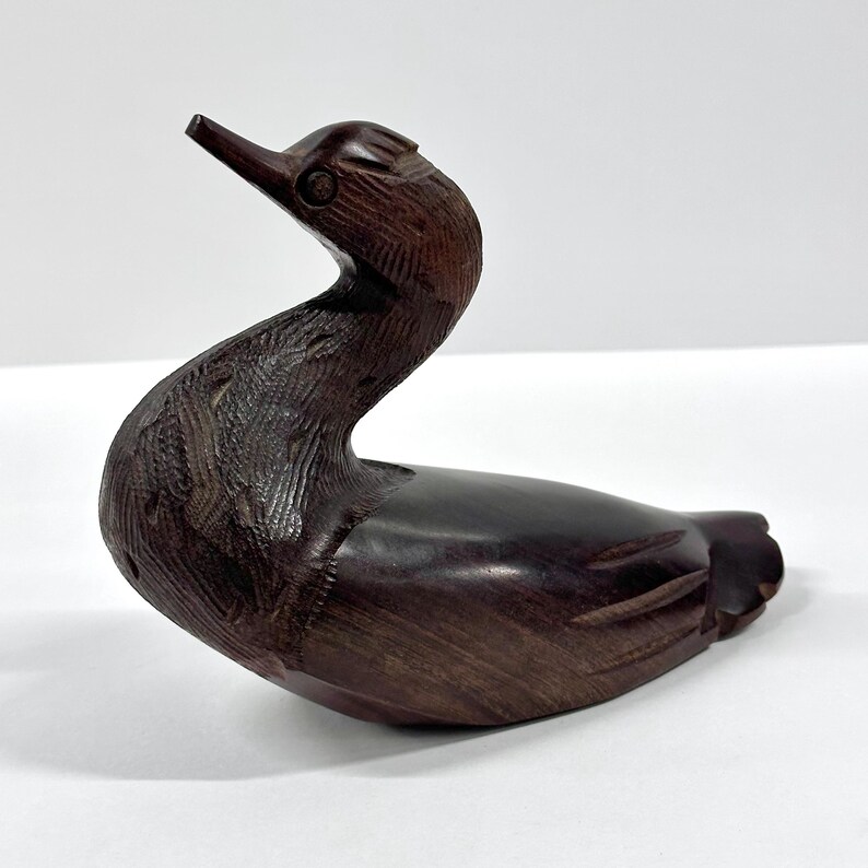 May include: A carved wooden duck figurine in a dark brown finish. The duck has a long, curved neck and detailed feather carvings. The beak is pointed upwards, and the duck is in a resting position, suggesting a decorative item or collectible.