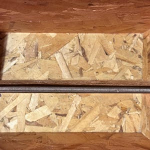 Op de afbeelding: Een close-up van de binnenkant van een houten kist. De kist is gemaakt van spaanplaat met een natuurlijke houtnerf afwerking. Een donkerbruine metalen staaf loopt horizontaal over het midden van de kist.