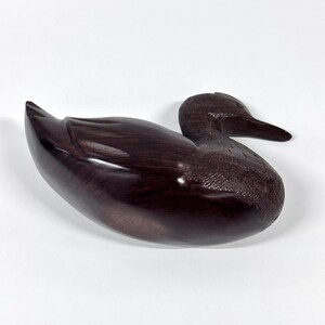 May include: A dark brown, wooden duck figurine. The duck is carved with smooth lines and a glossy finish. The duck is in a resting position, with its head turned to the side. The figurine is a decorative item.