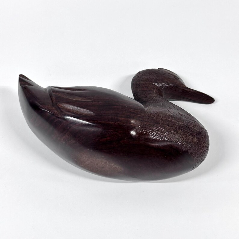 May include: A dark brown, wooden duck figurine. The duck is carved with smooth lines and a glossy finish. The duck is in a resting position, with its head turned to the side. The figurine is a decorative item.