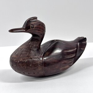 May include: A carved wooden duck figurine with a dark brown finish. The duck has a long beak, detailed feather carvings, and a smooth, glossy body. The figurine is a decorative item, likely for home decor.