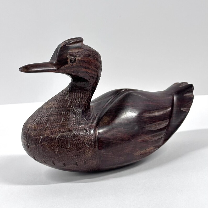 May include: A carved wooden duck figurine with a dark brown finish. The duck has a long beak, detailed feather carvings, and a smooth, glossy body. The figurine is a decorative item, likely for home decor.