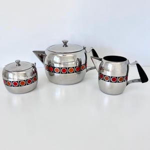 May include: A three-piece stainless steel tea set consisting of a teapot, a sugar bowl with a lid, and a creamer. Each piece features a horizontal band of alternating red and orange circles set within black squares against the polished metal surface.