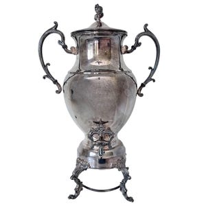 May include: A silver-plated beverage dispenser featuring a tall, bulbous body with two ornate, curved handles on either side. It includes a removable lid with a finial, a central spigot with decorative detailing, and stands on three scrolled legs.