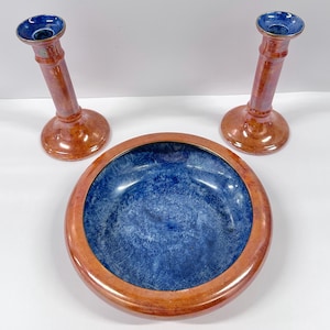 May include: A ceramic bowl and two candle holders. The bowl has a blue interior with a brown rim. The candle holders are brown with blue interiors. The items are arranged on a white surface.