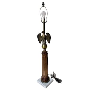 May include: A vintage table lamp featuring a brass eagle emblem. The lamp has a wooden column base, a marble square base, and a light bulb at the top. The lamp's cord is black and white.