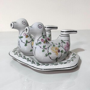 May include: A white ceramic duck-shaped salt and pepper shaker set with floral vine detailing. The set includes two duck shakers and a small tray, all with a black trim. The ducks have a whimsical design.