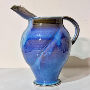 May include: A handmade ceramic pitcher with a blue glaze. The pitcher has a spout, a handle, and a wide base. The blue glaze has streaks of lighter blue and brown. The pitcher is a decorative item.