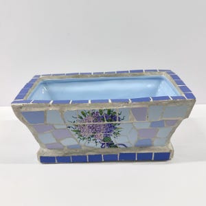 May include: Rectangular mosaic planter with a light blue interior. The exterior features a mosaic pattern of blue, lavender, and gray tiles. A floral bouquet design is on the front. The planter is suitable for indoor or outdoor use.