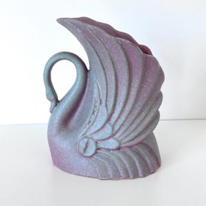 May include: A ceramic swan-shaped vase featuring a matte finish with a gradient of dusty purple and soft blue tones. The design includes stylized, raised wing feathers that curve upward, forming the opening of the vessel, with a curved neck.