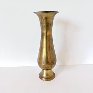 May include: A tall, slender brass vase with a polished metallic finish, featuring a flared trumpet-shaped opening at the top, a narrow neck, a rounded bulbous body, and a circular pedestal base that provides stability for the upright vessel.