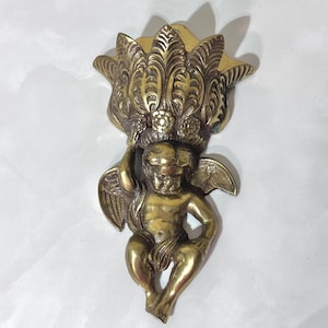 May include: A vintage brass wall sconce featuring a cherub holding a decorative floral element. The cherub has detailed wings and is in a seated position. The sconce has a warm, antique gold finish.