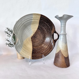 May include: A round ceramic serving platter with two side handles and a tall, slender ceramic vase. Both pieces feature a two-tone glaze in speckled light gray and dark brown, with visible concentric ridges and a textured, earthy finish.