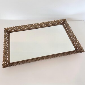 May include: A rectangular vanity tray featuring a central mirrored surface and an ornate, metallic gold-toned frame. The border is decorated with a continuous, raised leaf and vine pattern that extends around all four sides of the tray.