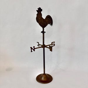 May include: A bronze-toned metal weather vane featuring a rooster silhouette mounted on a vertical rod. The structure includes a circular base and a directional crossbar with the letters N, S, E, and W clearly visible in a metallic finish.