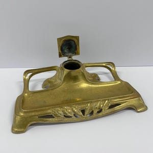 May include: Antique brass inkwell with a hinged lid and a decorative leaf design. The inkwell has a rectangular base with curved handles and a central ink pot. A vintage desk accessory.