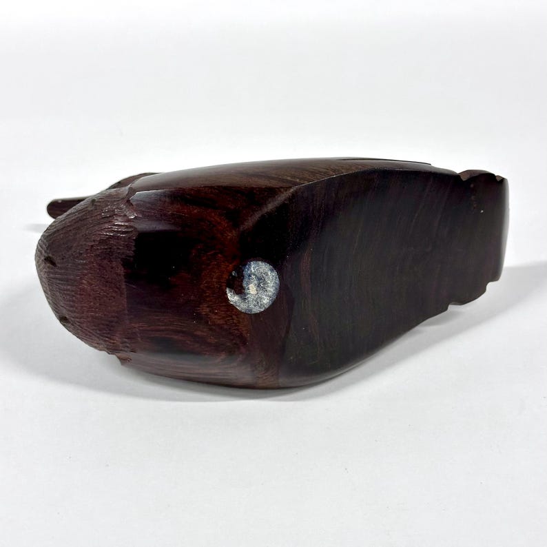 May include: A dark brown, carved wooden sculpture of a bird, possibly a duck or goose. The sculpture has a smooth, polished finish and a small, circular sticker on the underside. The bird's head is visible on the left side of the image.