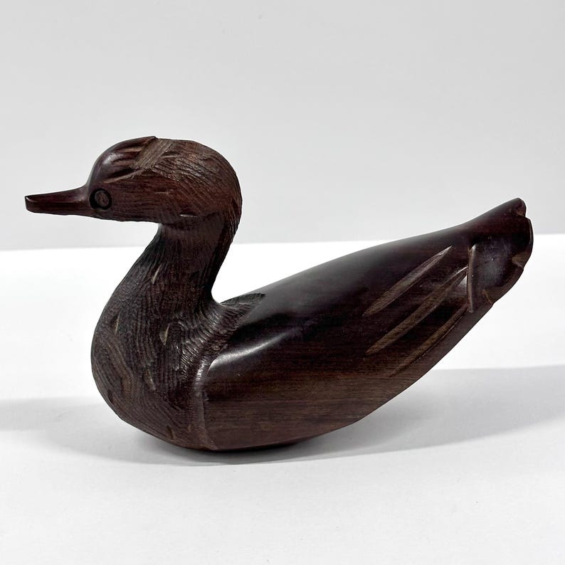 May include: A hand-carved wooden duck figurine. The duck is a rich, dark brown color with detailed carving on the head and body. The beak is pointed, and the duck is in a resting position.