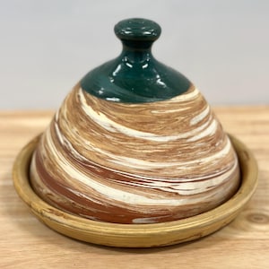 May include: A ceramic tagine pot with a conical lid and a round base. The lid is dark green, and the body features a swirl pattern of brown, white, and reddish-brown. The base is a light tan color. This traditional cookware is designed for slow-cooked stews.