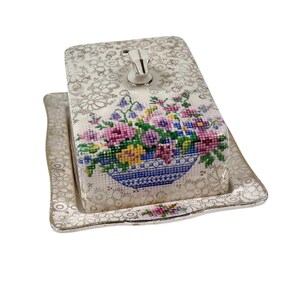 May include: A rectangular butter dish with a floral cross-stitch design on the lid. The lid features a silver handle and sits on a matching tray. The dish has a cream-colored background with gold floral accents.