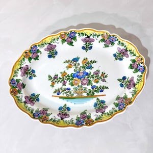 May include: Oval ceramic serving platter with a white base, adorned with a floral design. The rim is scalloped and outlined in yellow, with a repeating pattern of purple, blue, and yellow flowers. A central floral arrangement is also present.