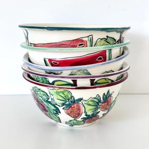 May include: A stack of four ceramic bowls with a cream-colored base. Each bowl features hand-painted illustrations of red strawberries and green leaves. The rims of the bowls are painted in varying colors including teal, light blue, and dark purple.